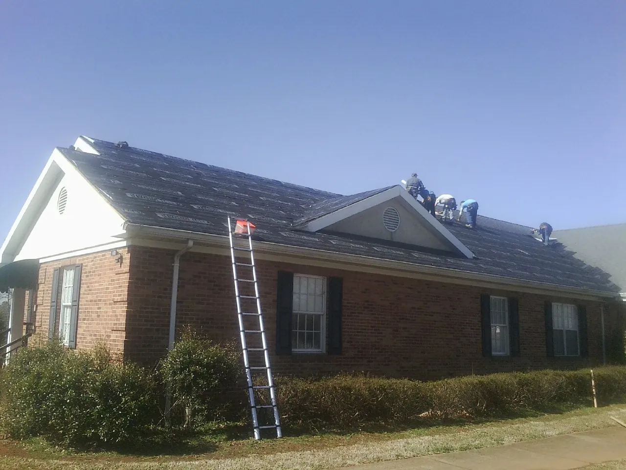 Crew tearing shingles off a brick ranch roof with a ladder set — Emergency Roof Repair work by our crew in Means