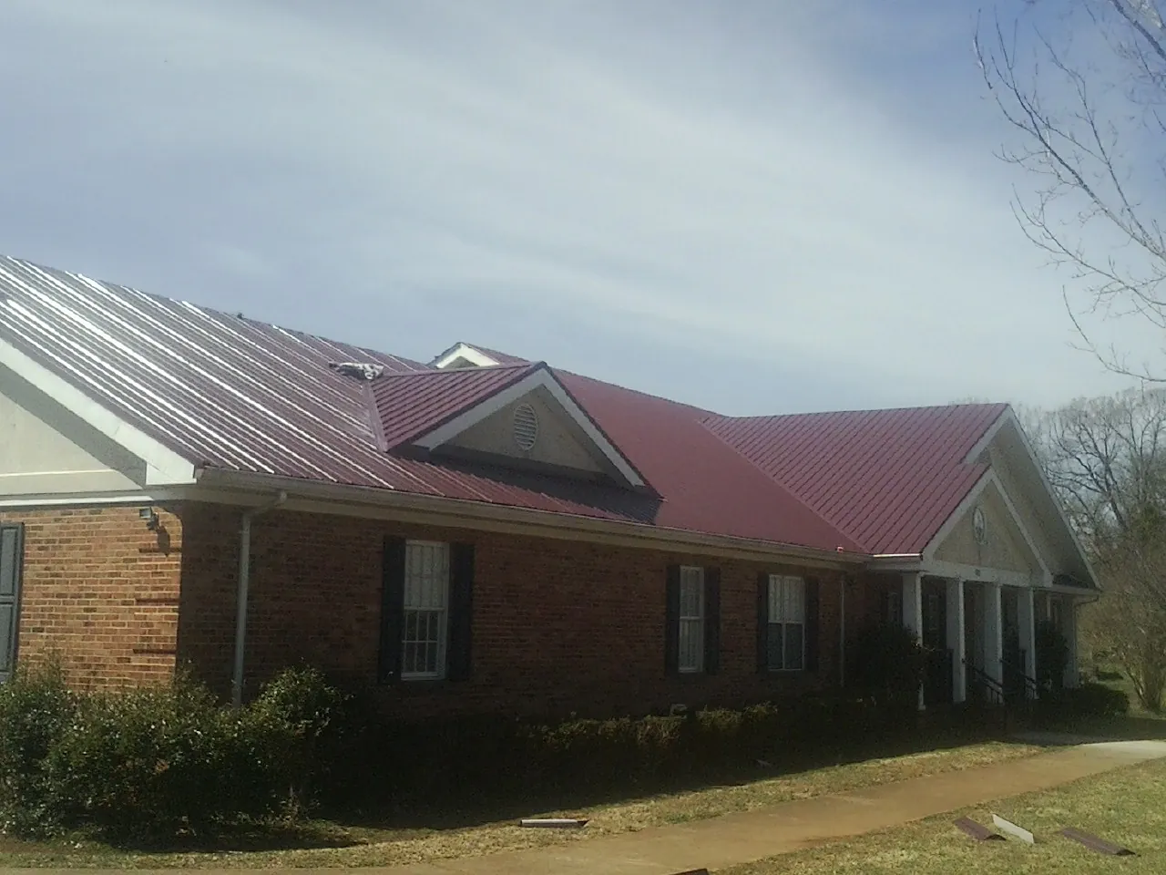 Red metal roof with gabled entries on a long brick building, from a Hail Damage Roof Repair job in Means